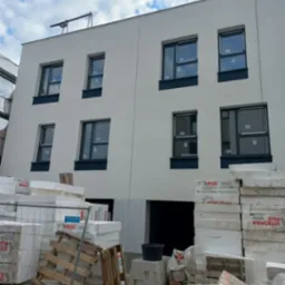 Ocieplony budynek z białymi ścianami i ciemnymi oknami. Na pierwszym planie styropian i materiały budowlane, sugerujące etap prac wykończeniowych elewacji.