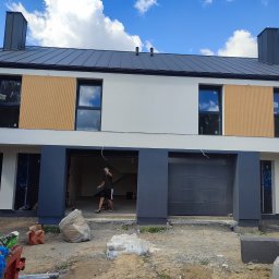 ONIX - Dwukondygnacyjny dom w trakcie prac elewacyjnych: białe ściany, szara stolarka okienna i fragmenty elewacji z drewnianymi panelami. Osoba w tle, materiały budowlane na pierwszym planie.