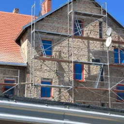 Budynek w rusztowaniach z anteną zbiorczą RTV-SAT na dachu, okna zabezpieczone folią. Widok na elewację podczas budowy instalacji.