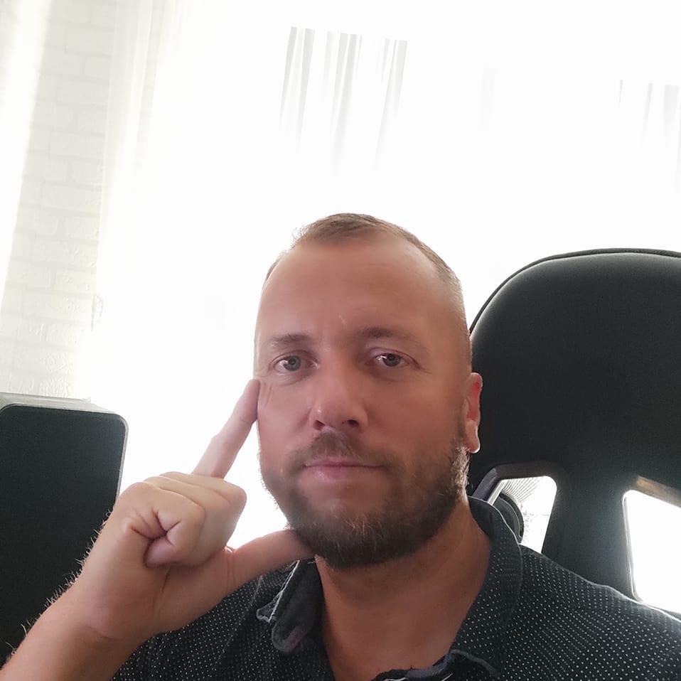 Portret brodatego mężczyzny w koszuli w kropki, siedzącego na czarnym krześle, z palcem wskazującym przy skroni i komputerem obok, na tle jasnego okna.