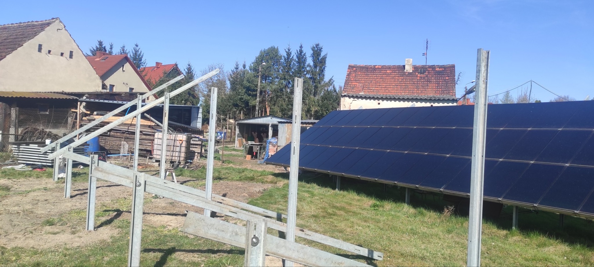 Montaż paneli fotowoltaicznych na trawiastym terenie, widoczne metalowe konstrukcje wsporcze, w tle zabudowania wiejskie i błękitne niebo. Częściowo ukończona instalacja.