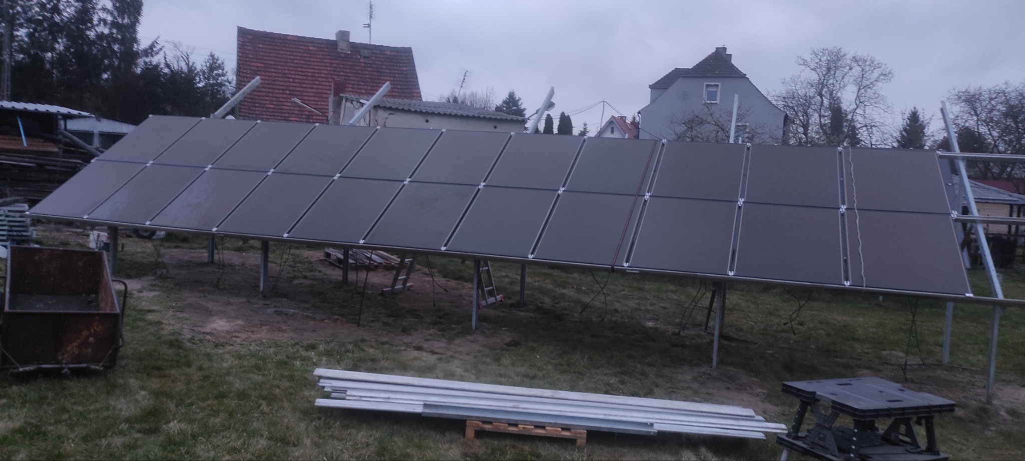 Montaż paneli słonecznych na metalowej konstrukcji w ogrodzie, widoczne narzędzia i materiały budowlane. Pochmurne niebo w tle.