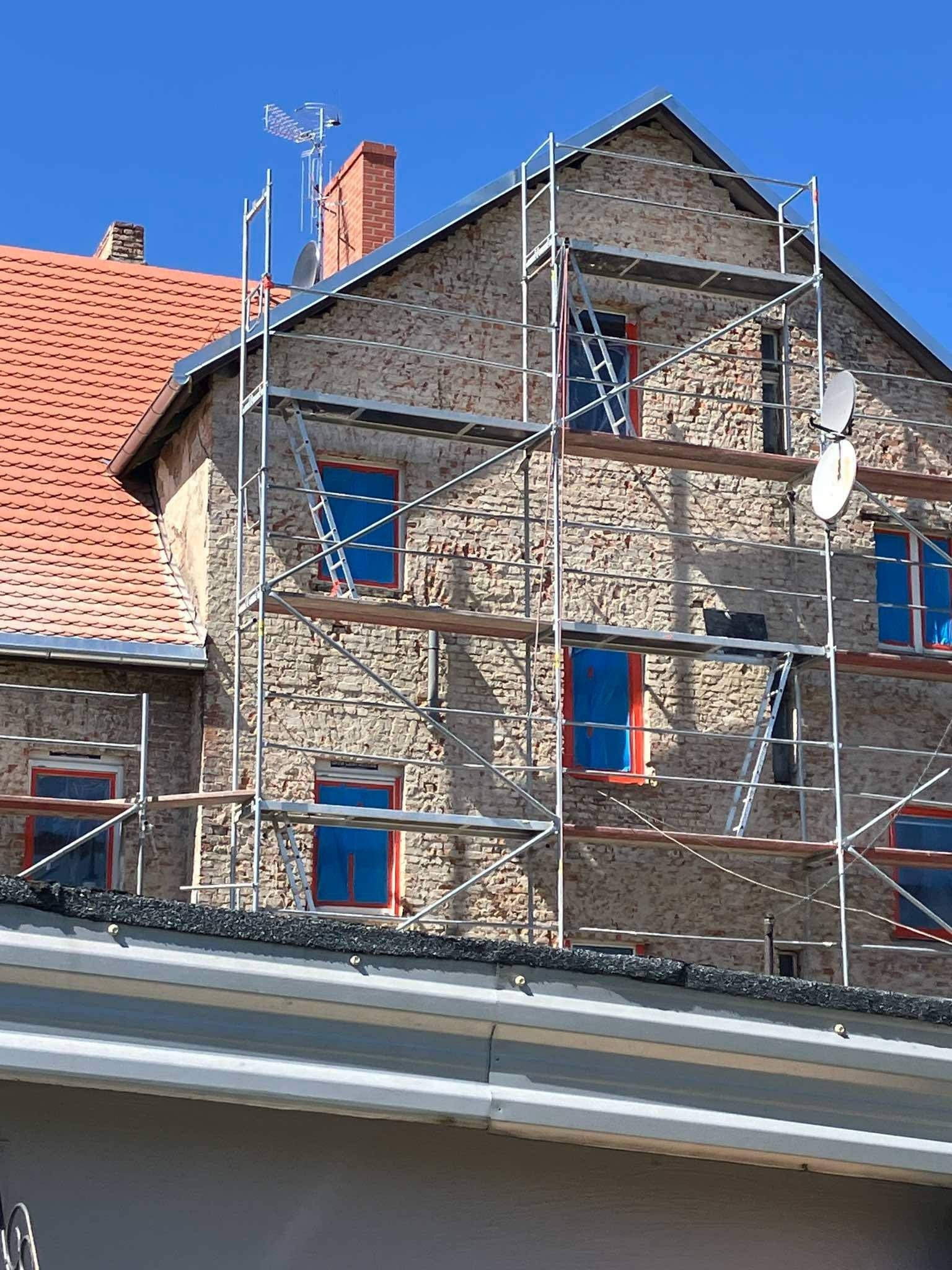 Budynek w rusztowaniach z anteną zbiorczą RTV-SAT na dachu, okna zabezpieczone folią. Widok na elewację podczas budowy instalacji.