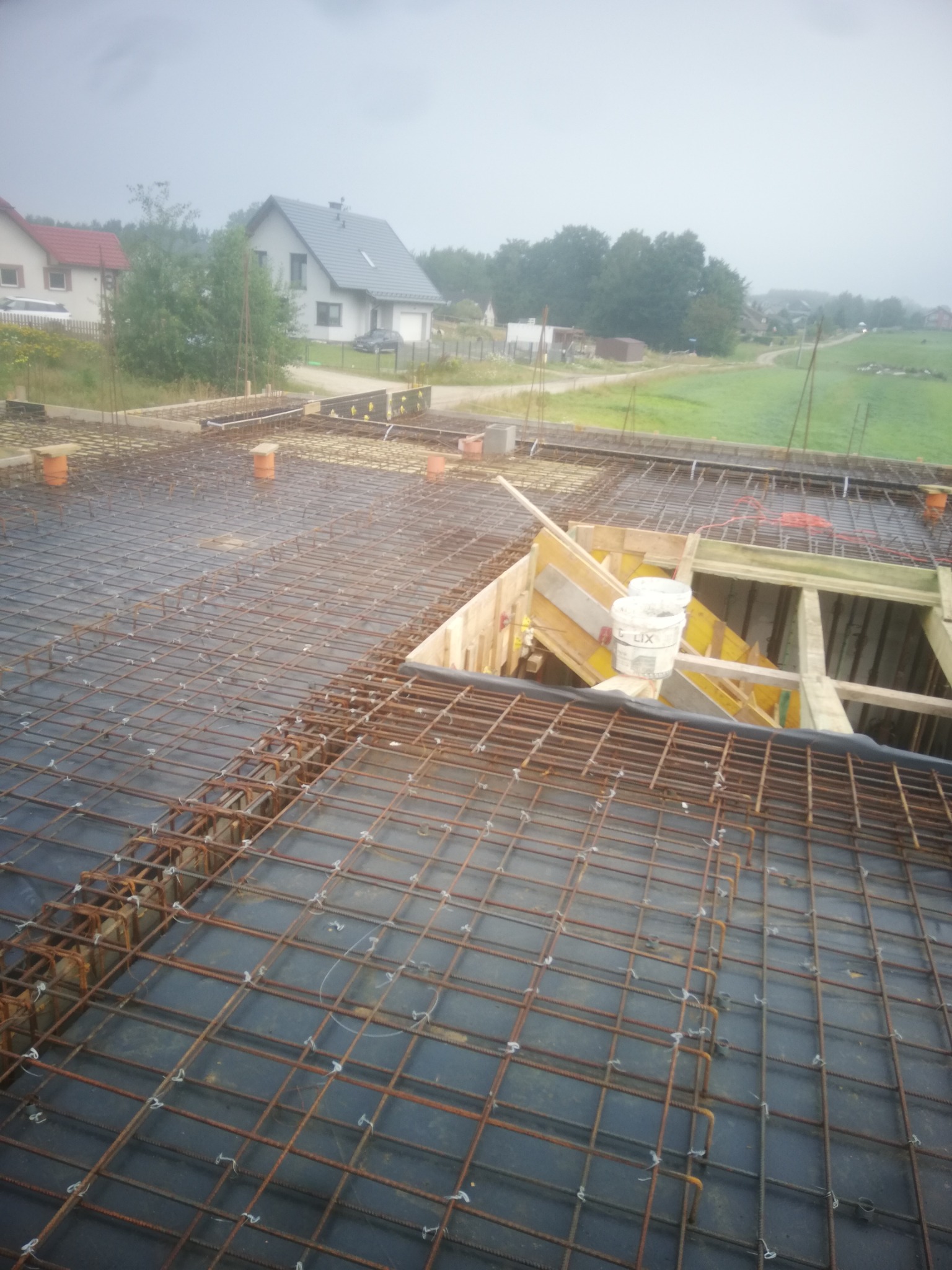 Zbrojenie stropu budynku jednorodzinnego z widocznymi elementami szalunku i kubełkami z zaprawą w tle, ujęcie z góry w pochmurny dzień.