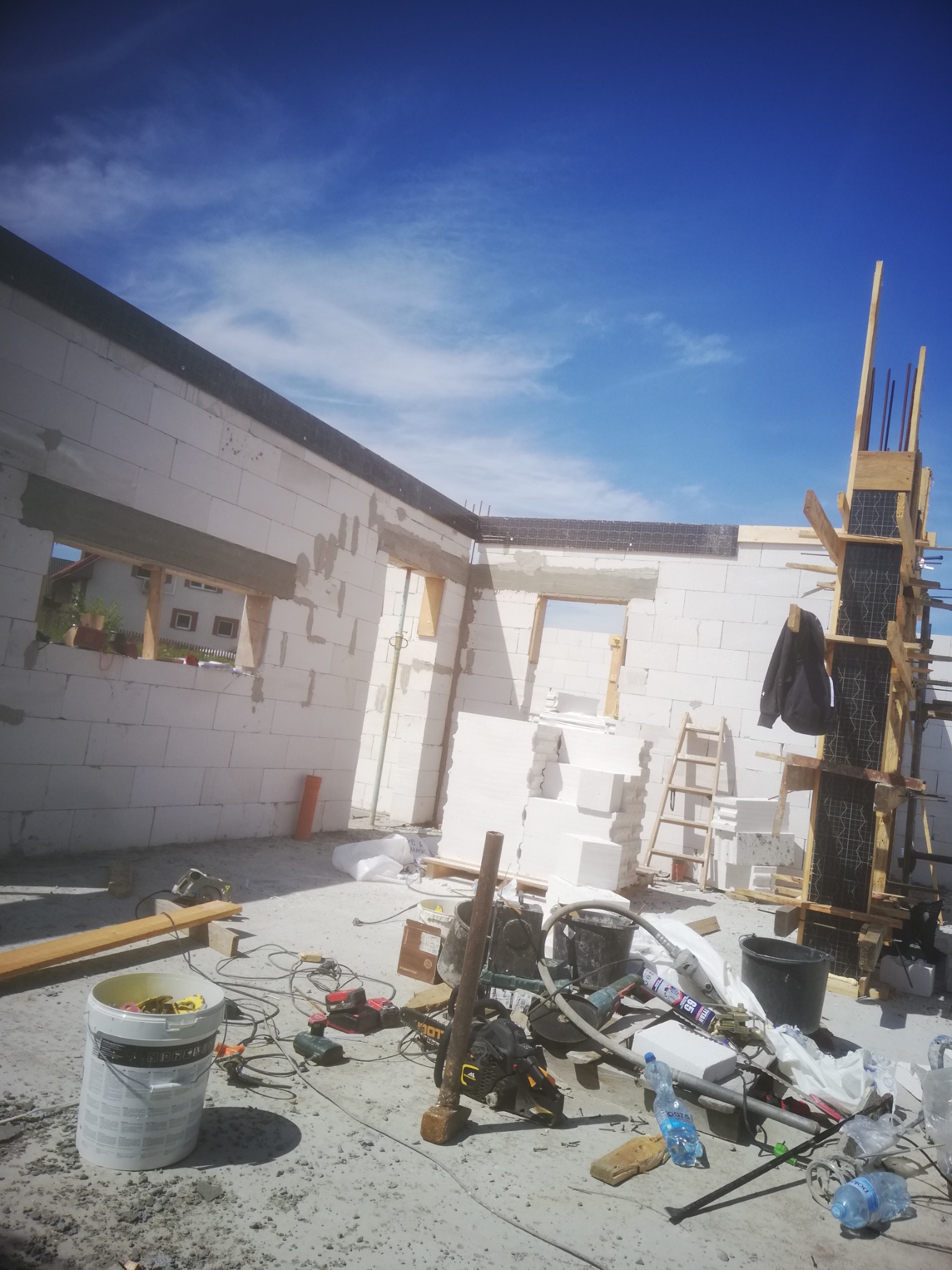 Budowa domu z białych bloczków: widoczne ściany, otwory okienne, szalunek kolumny. Na podłodze narzędzia i materiały budowlane. Niebo błękitne.