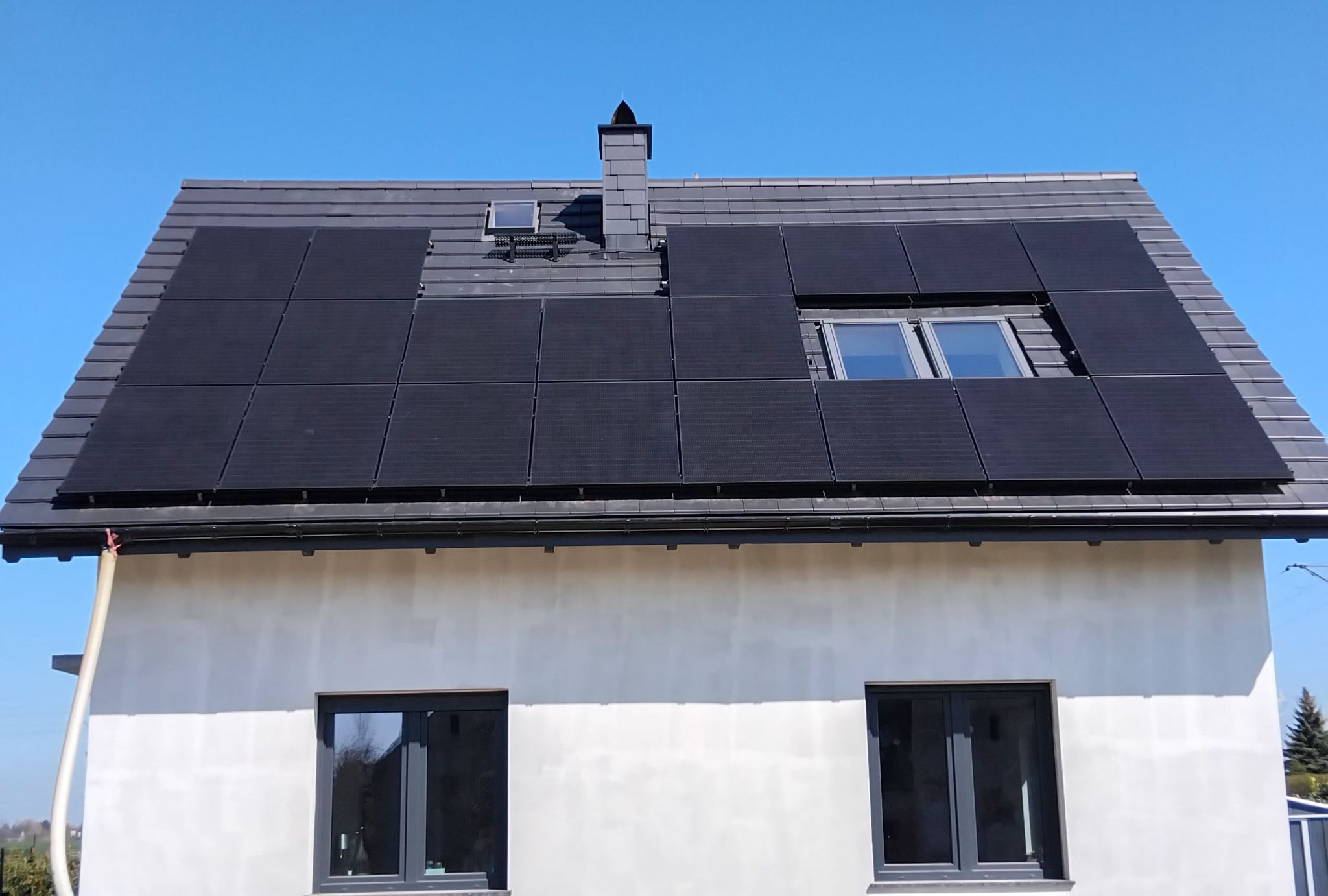 Ciemne panele fotowoltaiczne na szarym dachu domu jednorodzinnego z oknami dachowymi i kominem, na tle błękitnego nieba w słoneczny dzień.