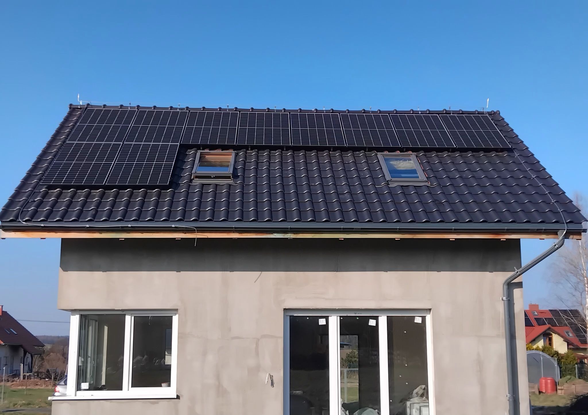 Dom jednorodzinny z panelami fotowoltaicznymi na ciemnym dachu, widoczne okna dachowe, jasne okna na parterze, surowy tynk na elewacji, błękitne niebo w tle.