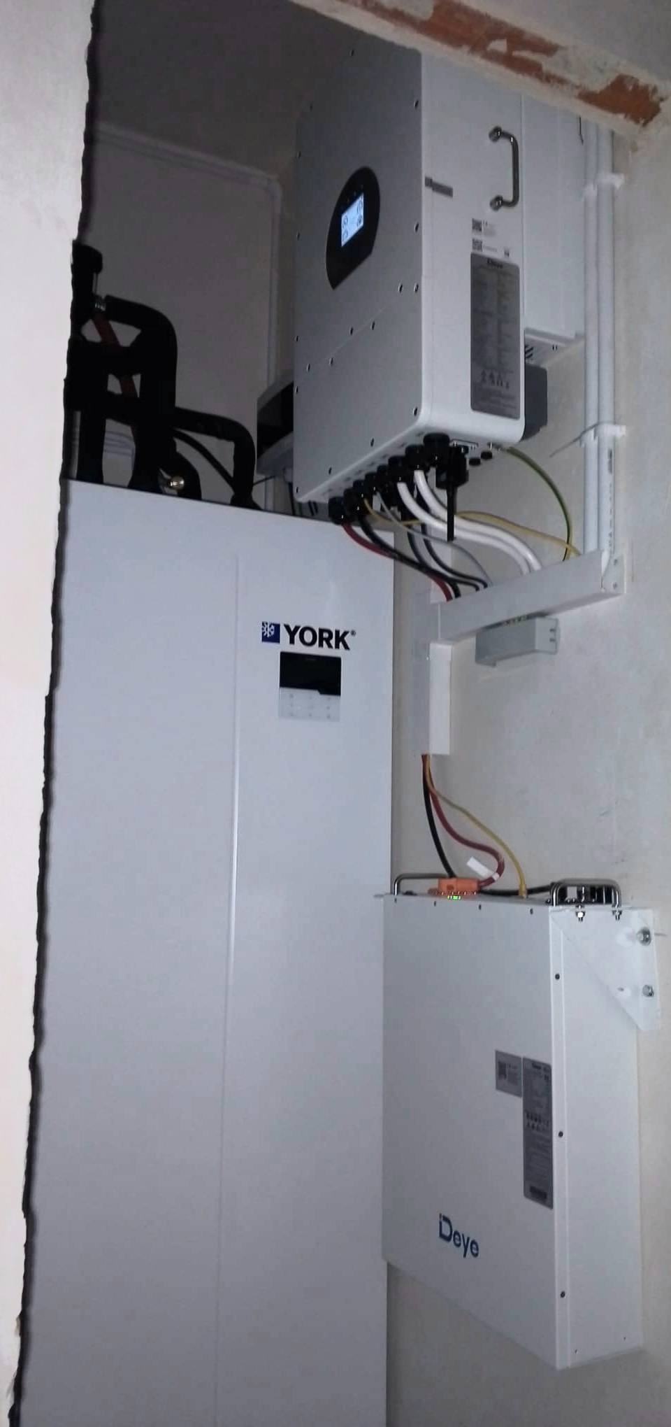 Zamontowana instalacja pompy ciepła YORK i falownika Deye z widocznymi przewodami w surowym wnętrzu budynku. Elementy instalacji przymocowane do ściany.