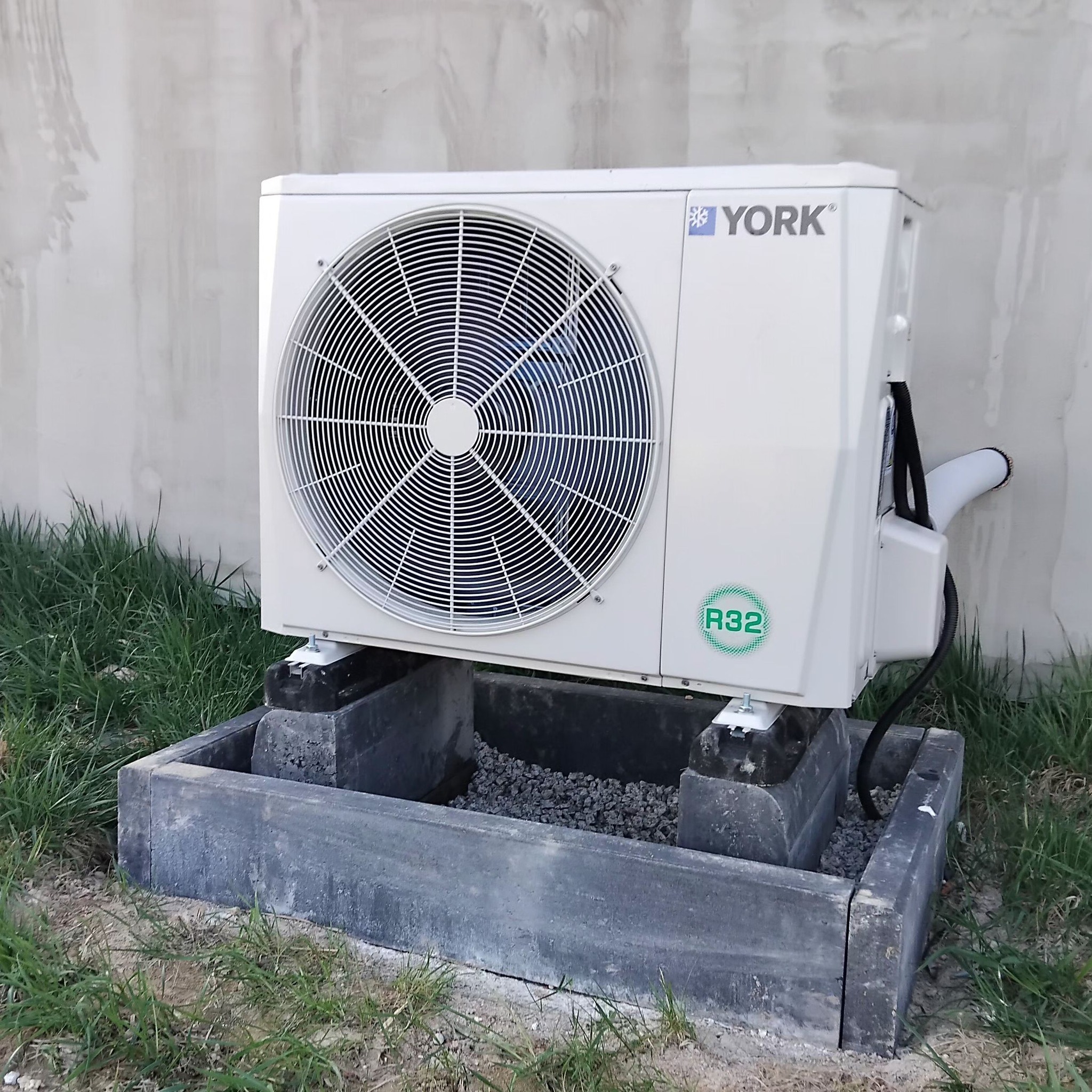 Biała zewnętrzna jednostka klimatyzacji YORK, zamontowana na betonowej podstawie z drobnymi kamieniami, na tle szarej ściany i trawy.