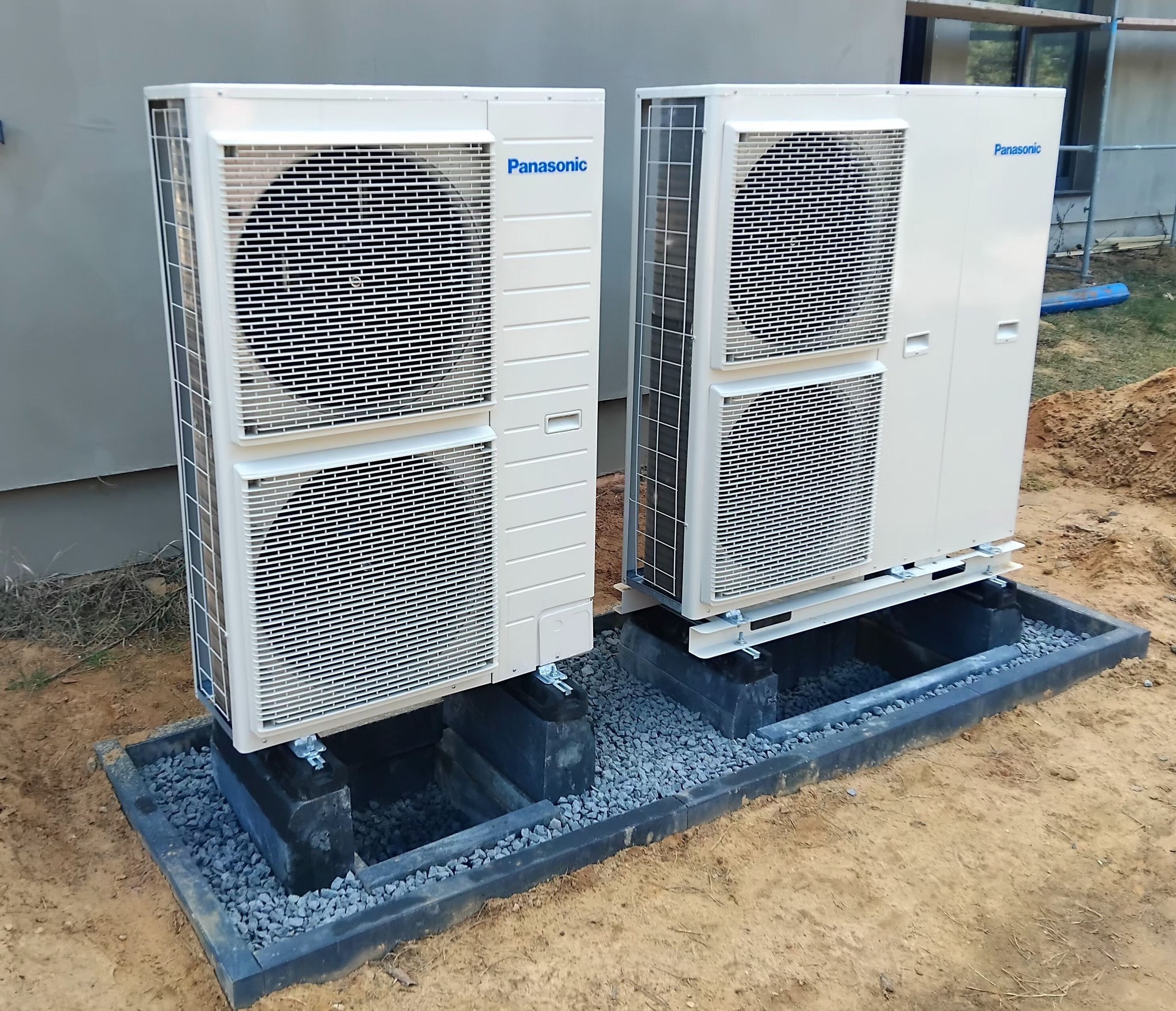 Dwie jednostki zewnętrzne klimatyzacji Panasonic, zamontowane na betonowych podstawach z podsypką z grysu, na zewnątrz budynku. Widoczne elementy instalacji.