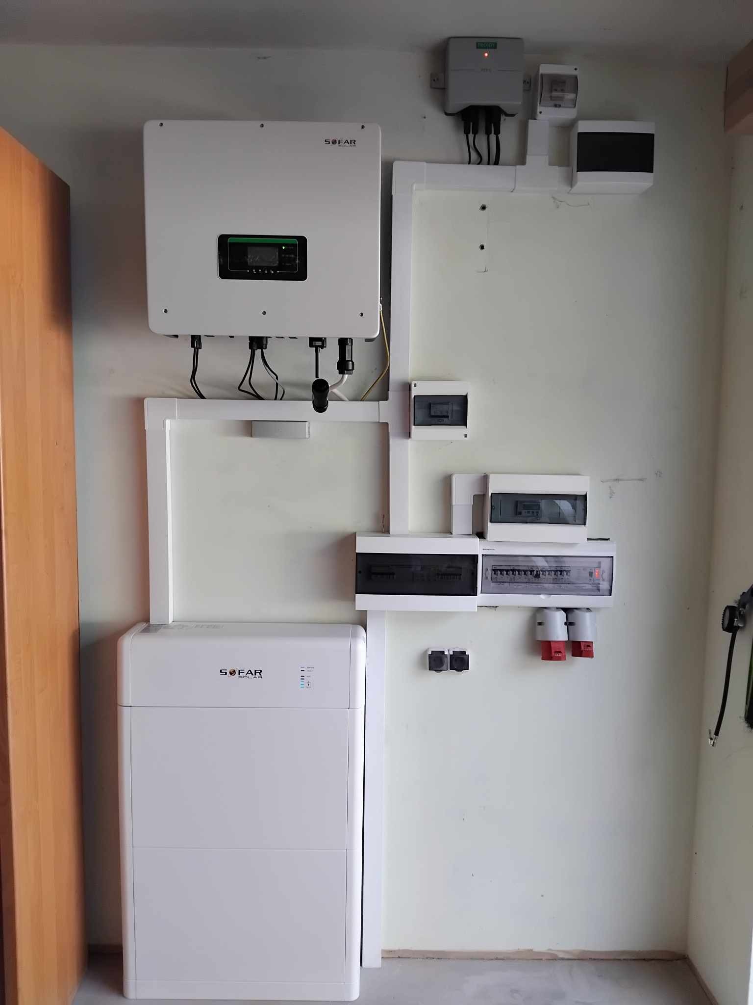 Kompletna instalacja fotowoltaiczna SOFAR z inwerterem, baterią i rozdzielnicą elektryczną na ścianie wewnątrz budynku. Widoczne białe obudowy urządzeń i przewody.