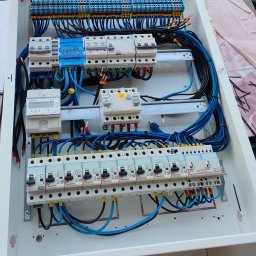 D.W Serwis Dariusz Zajdel - Tablica rozdzielcza z bezpiecznikami, przewodami w kolorach czarnym i niebieskim, oraz licznikiem energii elektrycznej. Widoczne oznaczenia i numery na elementach.