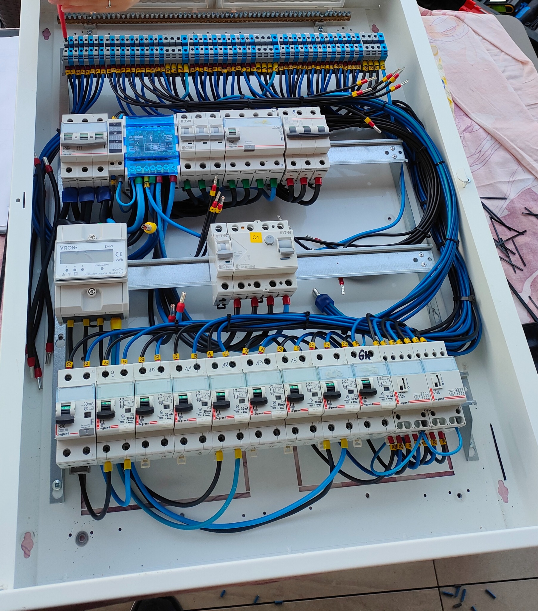 Tablica rozdzielcza z bezpiecznikami, przewodami w kolorach czarnym i niebieskim, oraz licznikiem energii elektrycznej. Widoczne oznaczenia i numery na elementach.