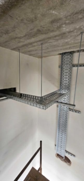 Metalowe korytka kablowe, zamocowane do sufitu i ściany. Elementy instalacji elektrycznej w surowym wnętrzu, widoczne z dołu. Szara betonowa powierzchnia sufitu.