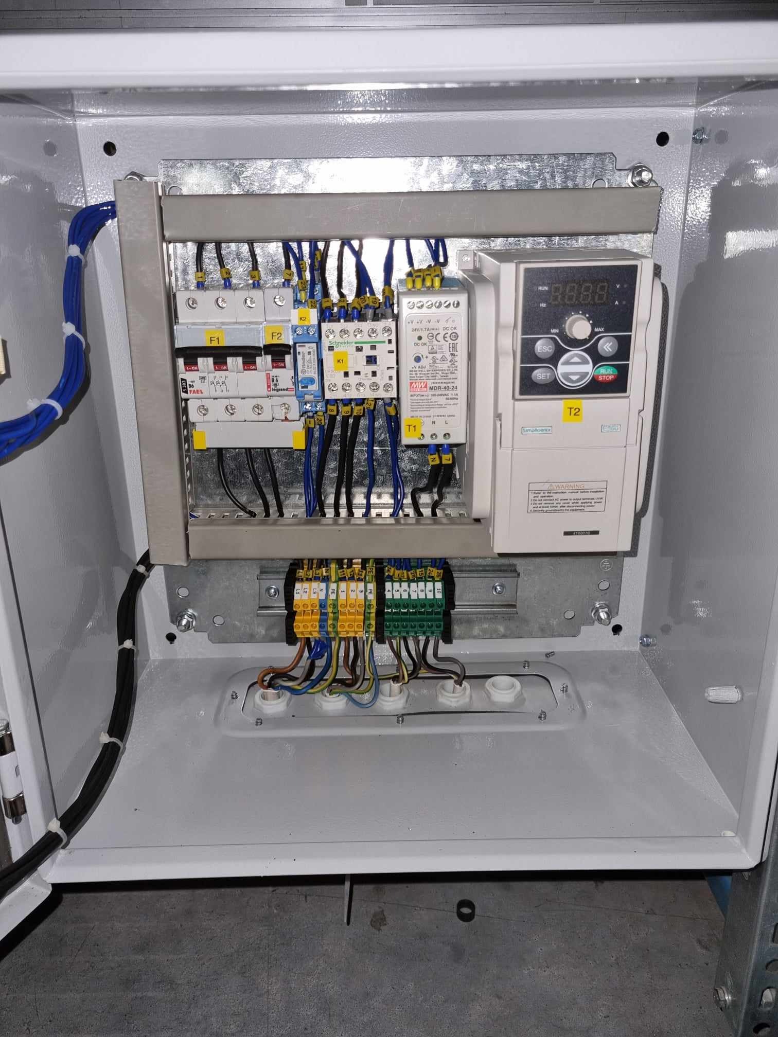 Wnętrze rozdzielnicy elektrycznej z bezpiecznikami, przewodami i sterownikiem, zamontowanymi na metalowej płycie w obudowie, widok z góry.
