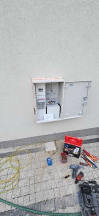 Otwarta, biała skrzynka elektryczna zamontowana na elewacji budynku. Obok, na kostce brukowej, leżą narzędzia elektryczne i zwój kabla.