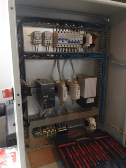 Otwarta szafa sterownicza z widocznymi elementami instalacji elektrycznej, takimi jak bezpieczniki, przekaźniki i zasilacz. W dolnej części skrzynka z narzędziami.