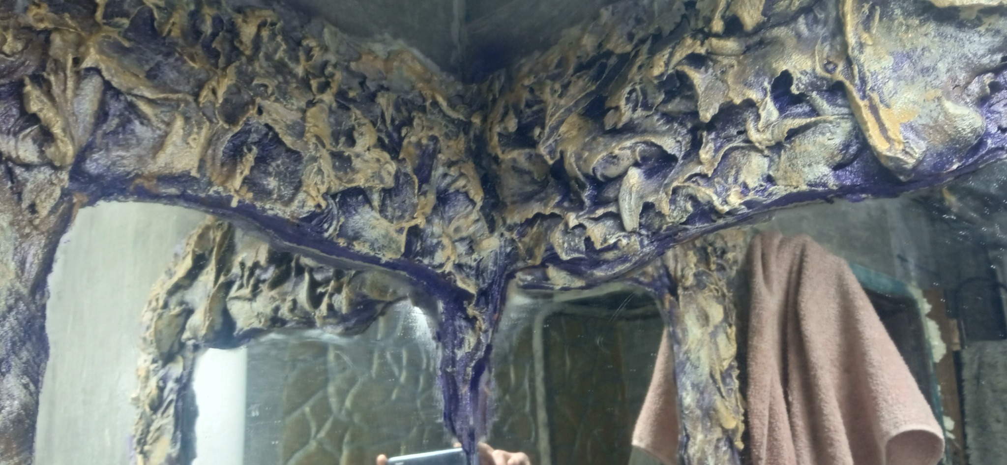 Dekoracyjna ściana z efektem kamienia w odcieniach złota i fioletu, odbijająca się w lustrze. Widoczny fragment ręcznika. Nietypowe wykończenie wnętrza.