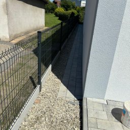 Patryk Kolasiński - Nowoczesne ogrodzenie panelowe, wąski chodnik z kostki, wysypane kamyki między ogrodzeniem a budynkiem. Widok z góry, słoneczny dzień, schludne wykonanie.
