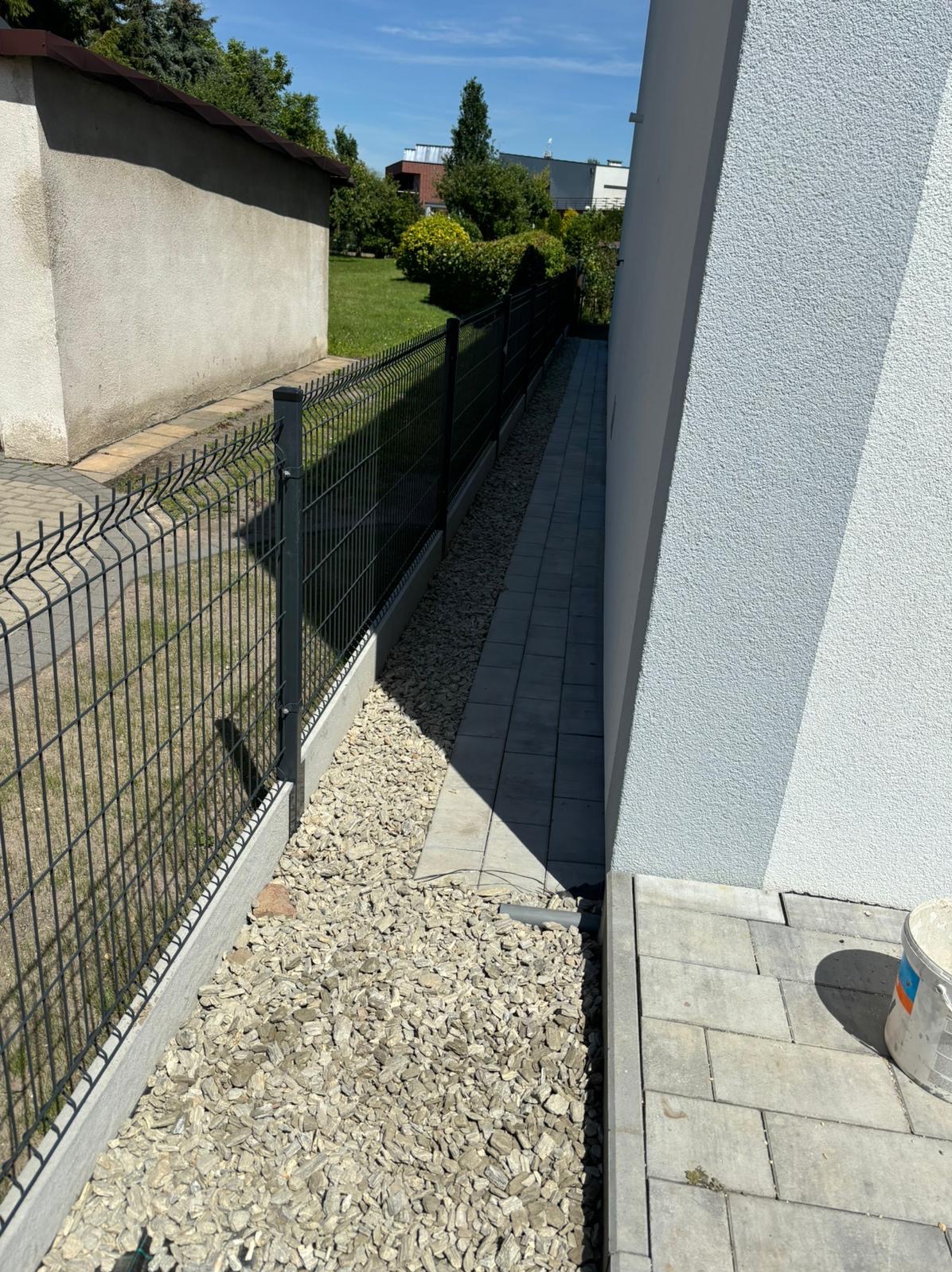 Nowoczesne ogrodzenie panelowe, wąski chodnik z kostki, wysypane kamyki między ogrodzeniem a budynkiem. Widok z góry, słoneczny dzień, schludne wykonanie.