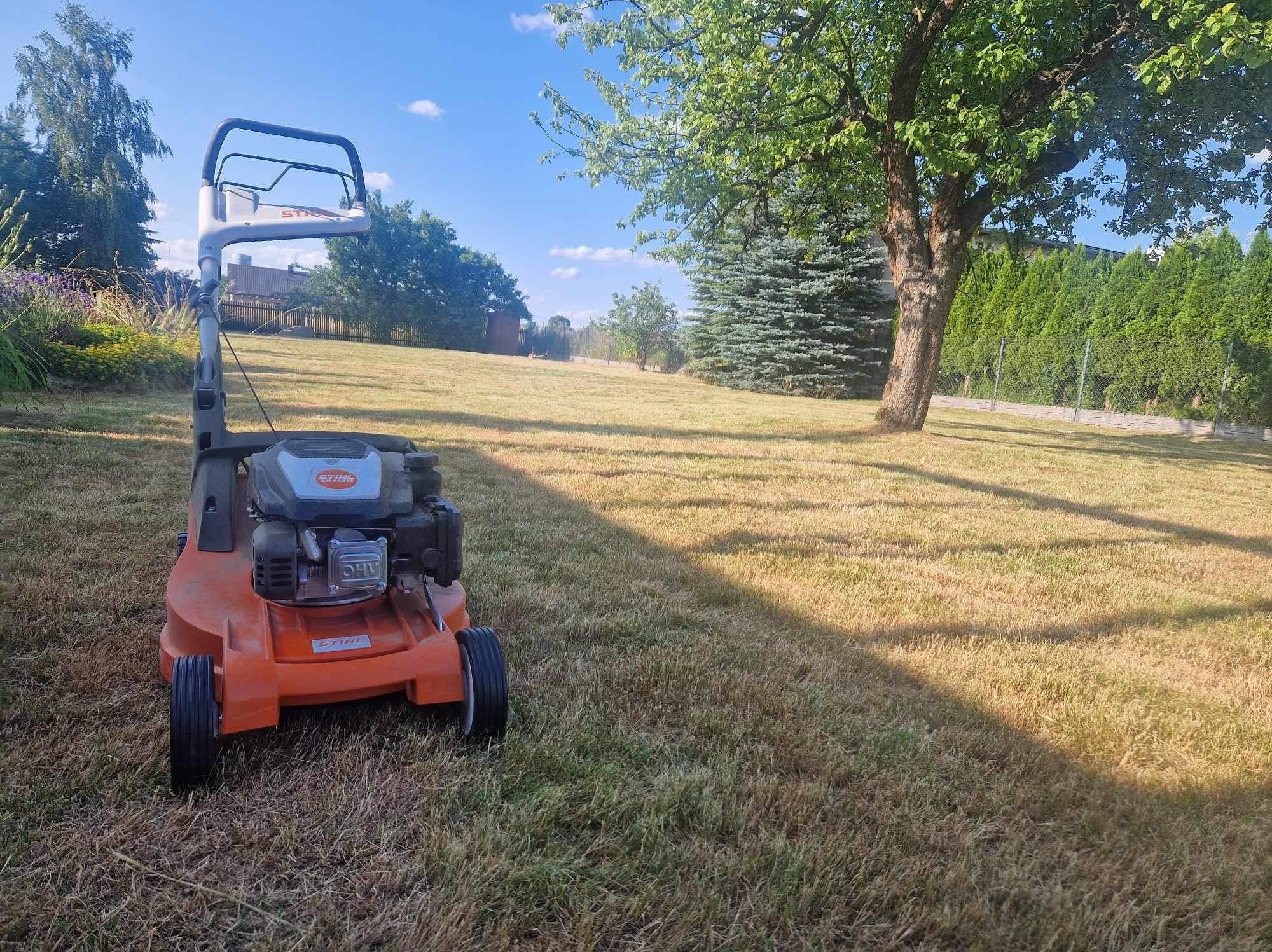 Pomarańczowa kosiarka spalinowa Stihl na tle wysuszonego trawnika przed domem z drzewami i żywopłotem, słoneczny dzień.