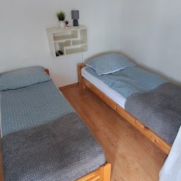ROYAL CLEANING ARTUR RUSZKOWSKI - Ustawione dwa pojedyncze łóżka z drewnianą ramą, przykryte szarą narzutą i niebieską pościelą. Na ścianie półka z dekoracjami. Na podłodze dywanik.