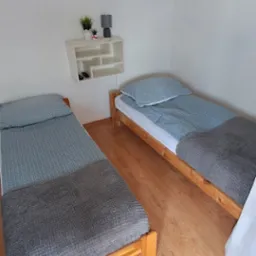 Ustawione dwa pojedyncze łóżka z drewnianą ramą, przykryte szarą narzutą i niebieską pościelą. Na ścianie półka z dekoracjami. Na podłodze dywanik.