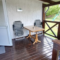 ROYAL CLEANING ARTUR RUSZKOWSKI - Zadaszony taras z drewnianą podłogą po czyszczeniu i olejowaniu. Dwa krzesła i stół na werandzie. W tle widoczny plac zabaw.