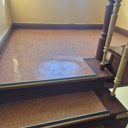 ROYAL CLEANING ARTUR RUSZKOWSKI - Wyczyszczona podłoga na podeście schodowym z drewnianą balustradą, widoczne ślady po umyciu. Żółte ściany i okno w tle. Ujęcie z dołu, z perspektywy schodów.