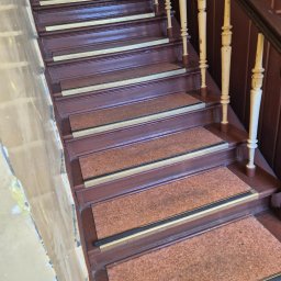 ROYAL CLEANING ARTUR RUSZKOWSKI - Schody drewniane z brązowymi stopniami i beżowymi balustradami. Na stopniach nakładki z korka z metalowymi listwami. Ściana obok schodów z widocznymi śladami po remoncie.