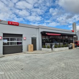 ROYAL CLEANING ARTUR RUSZKOWSKI - Nowoczesna, szara myjnia samochodowa z czerwonymi akcentami i napisem 'Myjnia'. Widoczne bramy wjazdowe, okna kawiarni 'Stop Cafe' i kostka brukowa na placu.