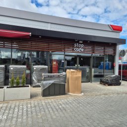 ROYAL CLEANING ARTUR RUSZKOWSKI - Nowe nasadzenia i elementy małej architektury przed lokalem Stop Cafe, zapakowane w folię i karton, czekają na montaż. Widoczny czerwony van dostawczy.