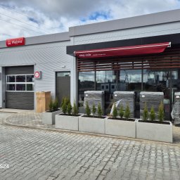 ROYAL CLEANING ARTUR RUSZKOWSKI - Nowoczesna myjnia samochodowa z kawiarnią Stop Cafe, ozdobiona rzędem betonowych donic z młodymi tujami. Kostka brukowa na pierwszym planie.