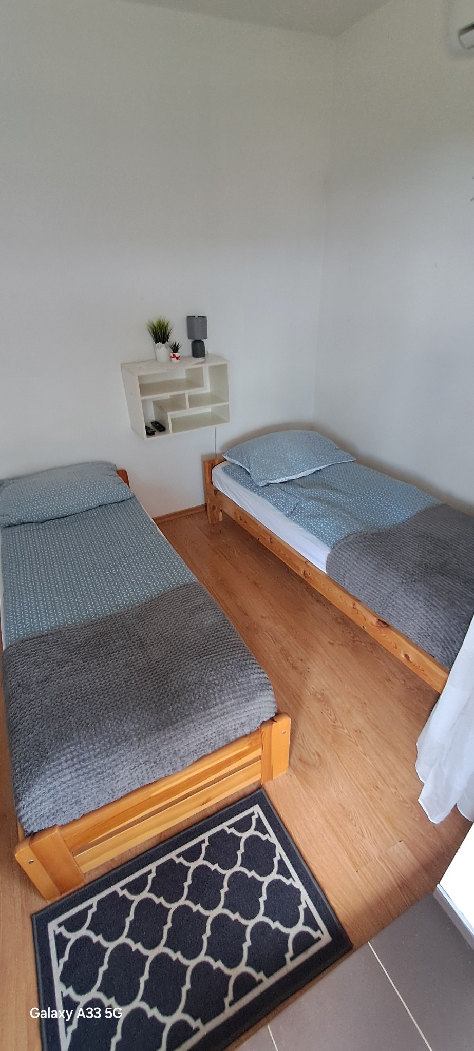 Ustawione dwa pojedyncze łóżka z drewnianą ramą, przykryte szarą narzutą i niebieską pościelą. Na ścianie półka z dekoracjami. Na podłodze dywanik.