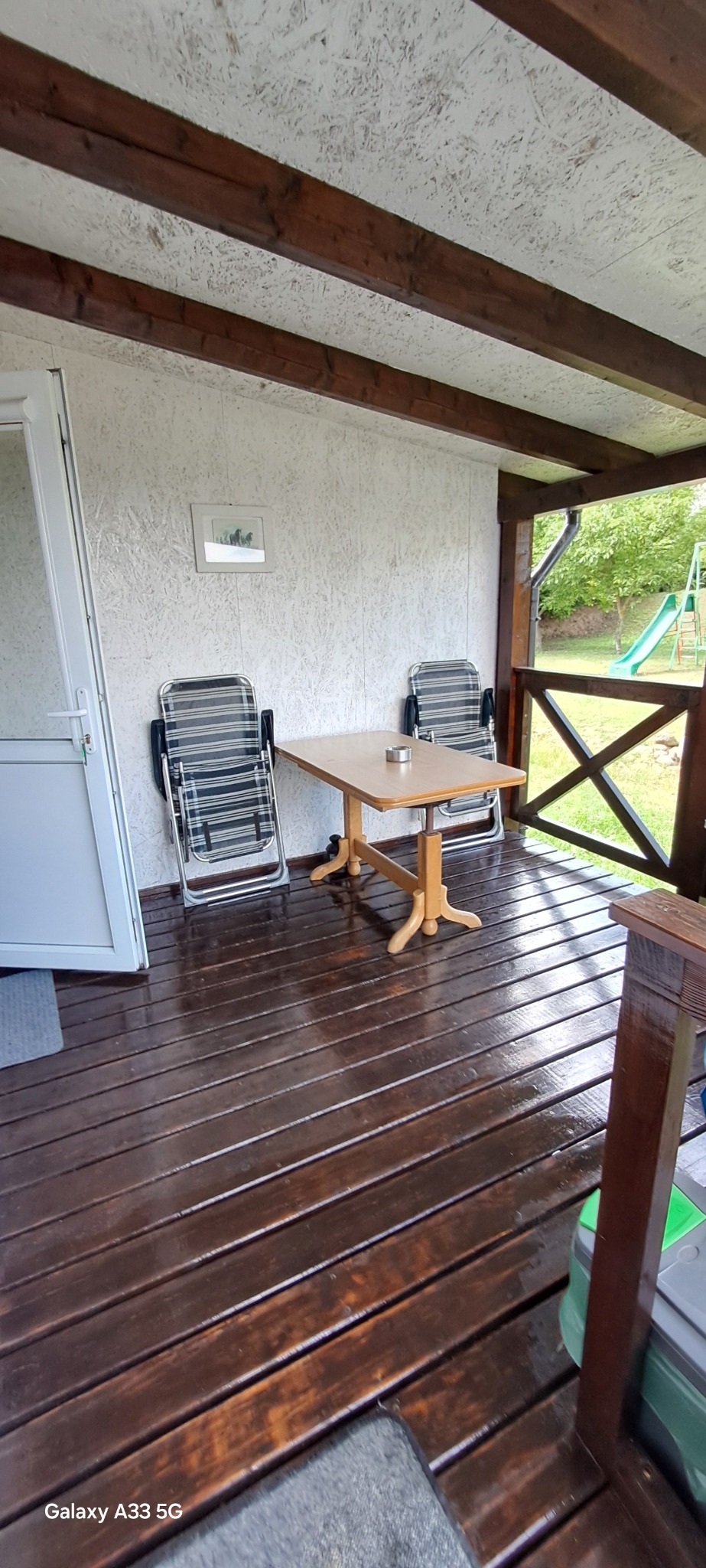 Zadaszony taras z drewnianą podłogą po czyszczeniu i olejowaniu. Dwa krzesła i stół na werandzie. W tle widoczny plac zabaw.