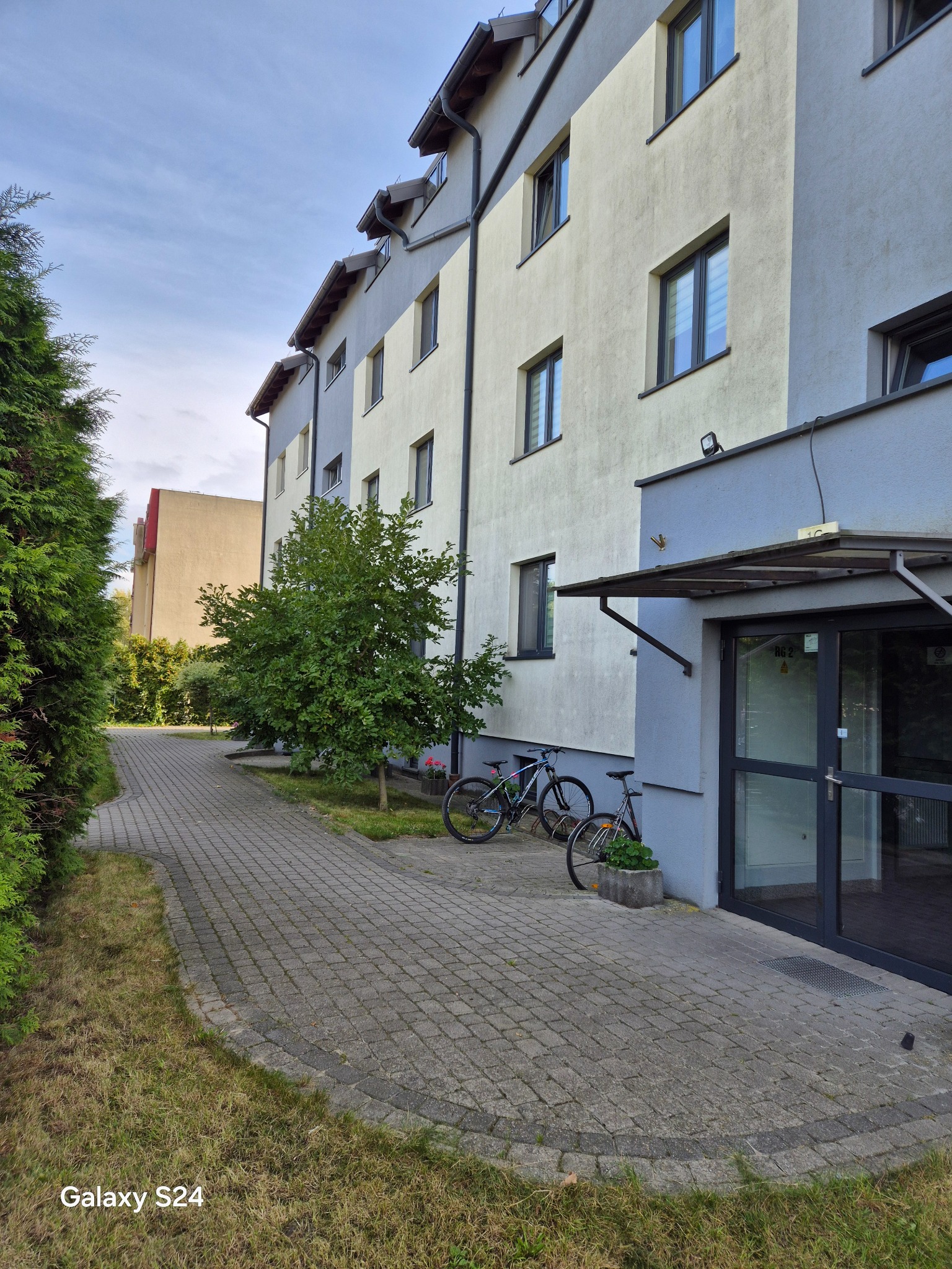 Dwupiętrowy budynek z szarą elewacją, widok z chodnika z kostki brukowej. Dwa rowery zaparkowane przy wejściu. Z prawej strony drzwi wejściowe.