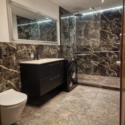 Firma ogólnobudowlana ariel Kowalke - Elegancka łazienka z czarnymi płytkami imitującymi marmur, kabiną prysznicową typu walk-in, umywalką z szafką i toaletą. Obok umywalki stoi pralka.