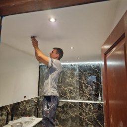 Firma ogólnobudowlana ariel Kowalke - Elektryk montuje oświetlenie w łazience z marmurowymi płytkami. Widoczne narzędzia i elementy instalacji. Biała sufit i częściowo wykończone ściany.