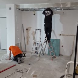 Firma ogólnobudowlana ariel Kowalke - Prace wykończeniowe w surowym wnętrzu: montaż sufitu podwieszanego i instalacji elektrycznej. Dwóch pracowników na drabinie i na podłodze. Widoczne narzędzia i materiały.