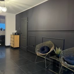 Firma ogólnobudowlana ariel Kowalke - Czarne płytki podłogowe w salonie, ściana z lameli w kolorze grafitowym, złote krzesła z poduszkami, drewniana komoda i drzwi w odcieniach szarości.