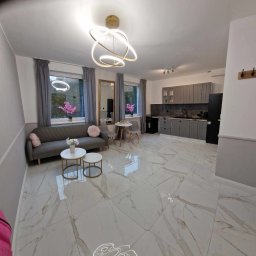 Firma ogólnobudowlana ariel Kowalke - Jasne, przestronne wnętrze z białymi marmurowymi płytkami podłogowymi. Szara sofa, okna z zasłonami i kuchnia w tle. Nowoczesna lampa sufitowa.