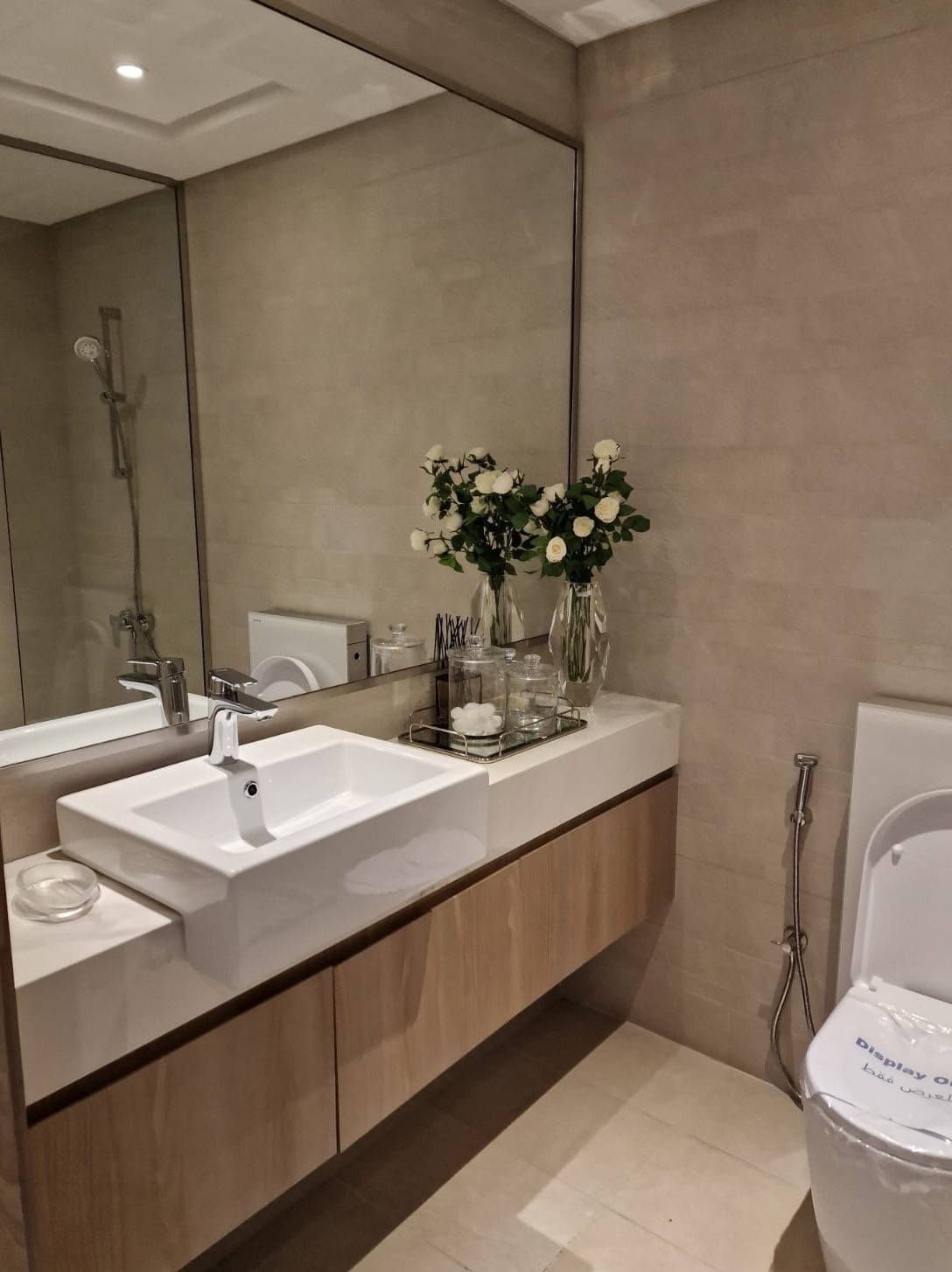 Elegancka łazienka z umywalką nablatową, lustrem w ramie i dekoracyjnymi wazonami z białymi różami. Nowoczesne wykończenie, beżowe płytki, czysta armatura.