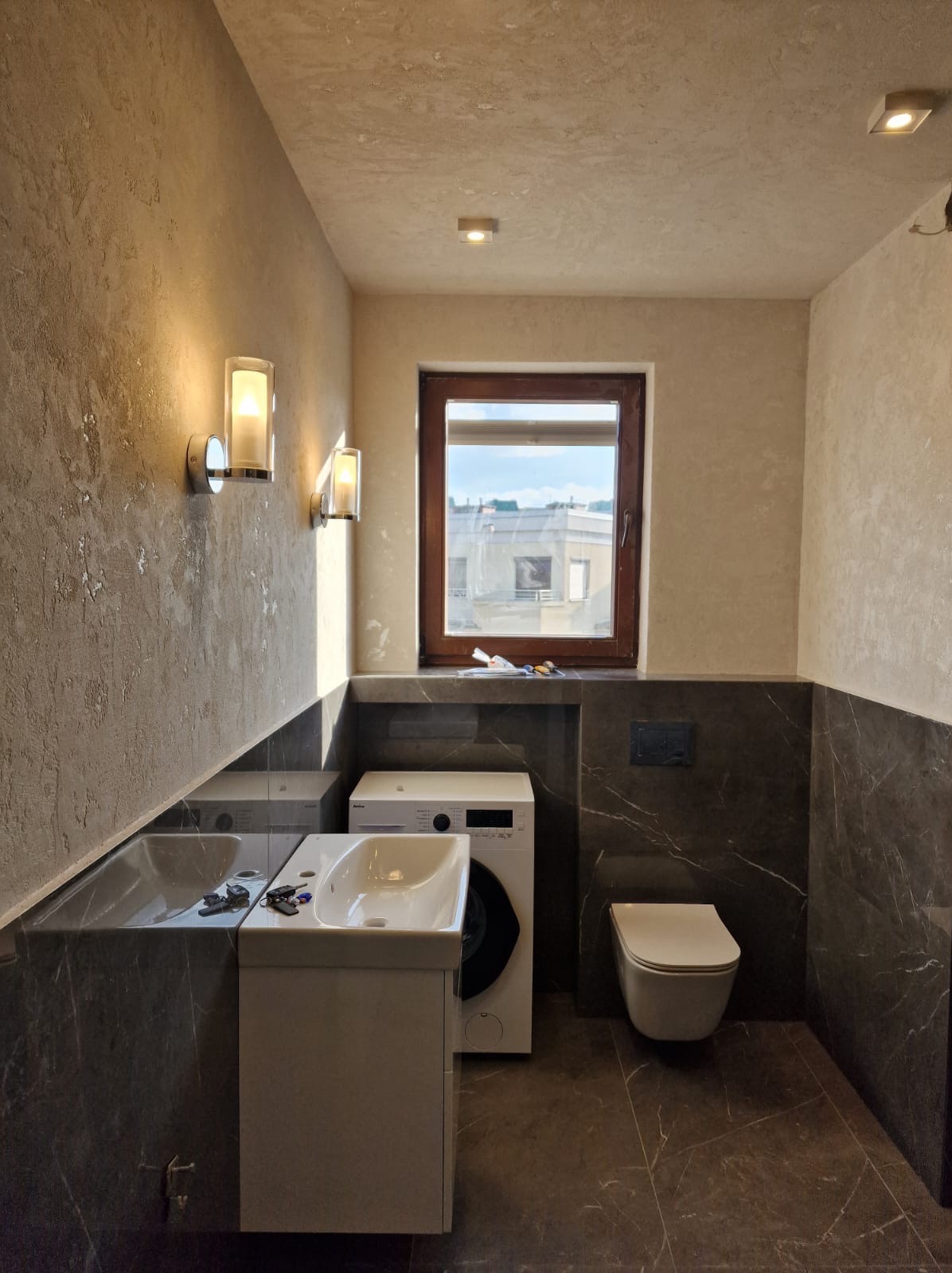 Nowoczesna łazienka z dwoma umywalkami, pralką i toaletą w kolorach szarości i beżu, oświetlona kinkietami i światłem dziennym z okna.