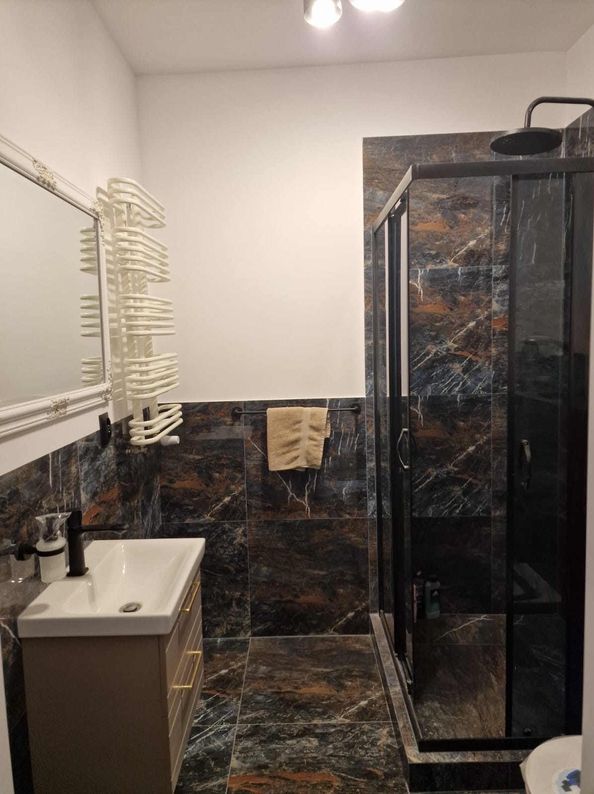 Wykończona łazienka w Skarszewach z kabiną prysznicową w czarnej ramie, umywalką z szafką i grzejnikiem drabinkowym na tle płytek imitujących marmur.