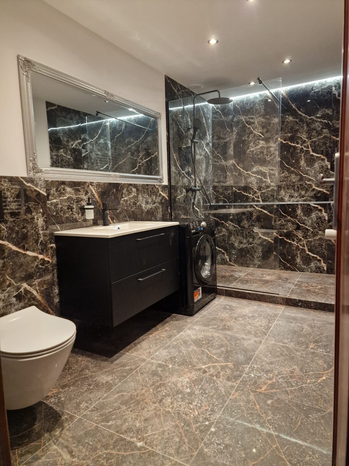 Elegancka łazienka z czarnymi płytkami imitującymi marmur, kabiną prysznicową typu walk-in, umywalką z szafką i toaletą. Obok umywalki stoi pralka.