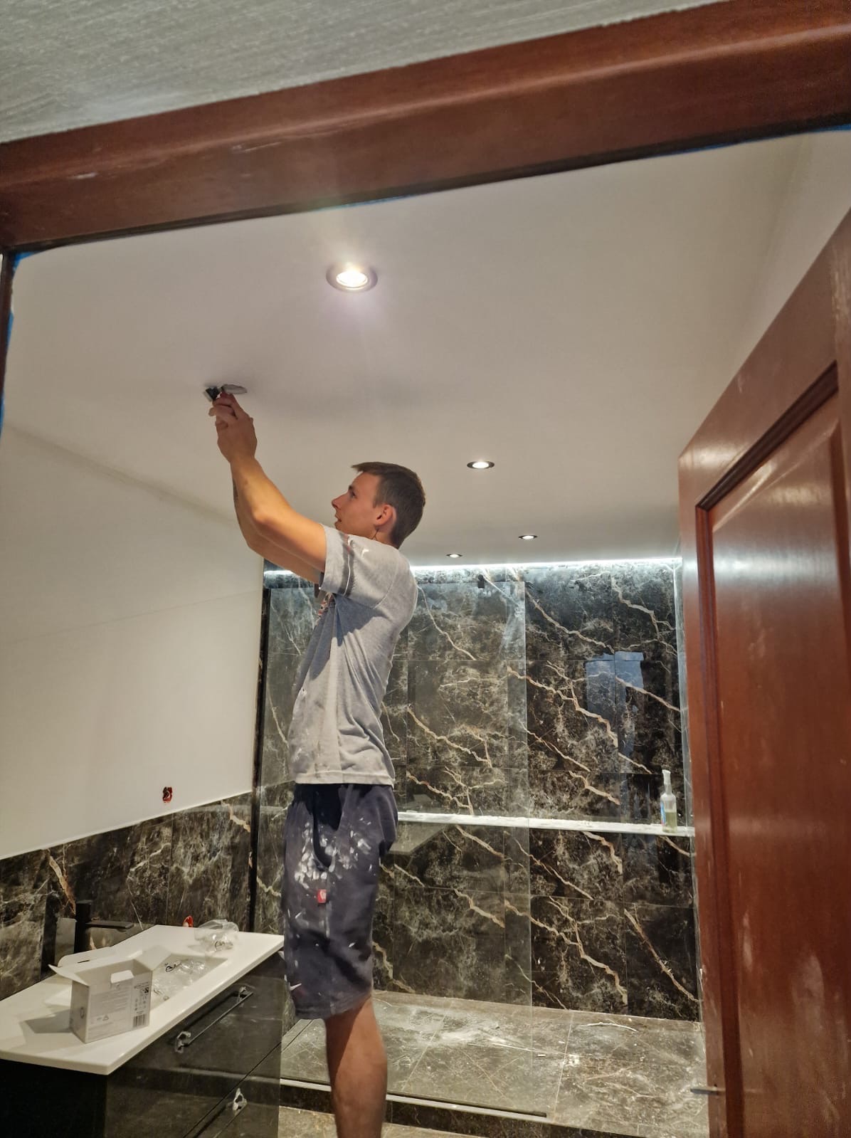 Elektryk montuje oświetlenie w łazience z marmurowymi płytkami. Widoczne narzędzia i elementy instalacji. Biała sufit i częściowo wykończone ściany.