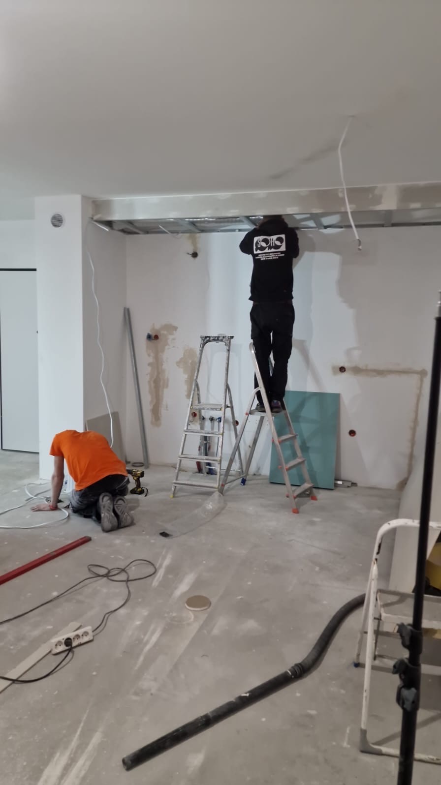 Prace wykończeniowe w surowym wnętrzu: montaż sufitu podwieszanego i instalacji elektrycznej. Dwóch pracowników na drabinie i na podłodze. Widoczne narzędzia i materiały.