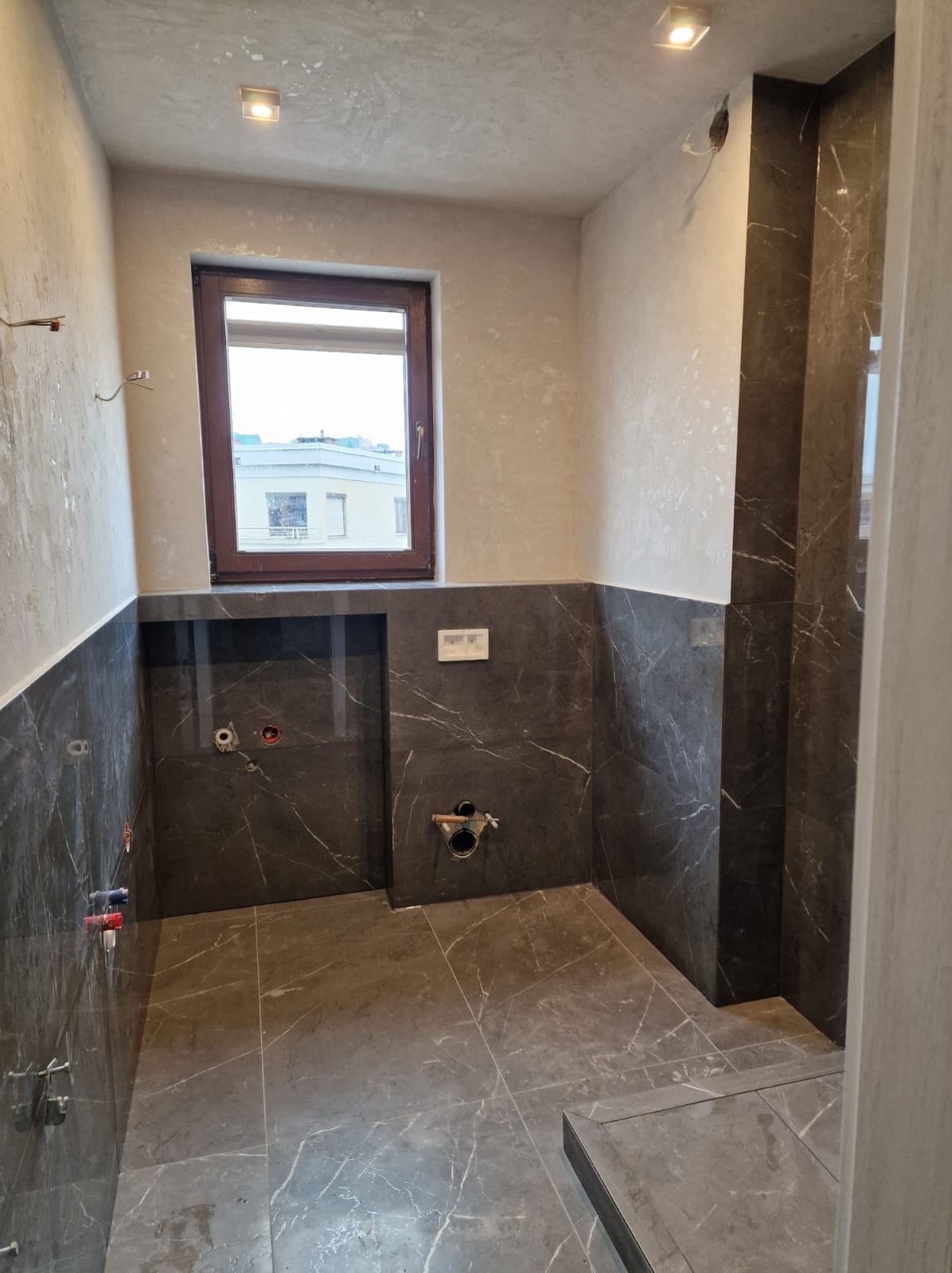 Łazienka w trakcie remontu: szare płytki imitujące marmur na podłodze i ścianach do połowy wysokości, widoczne instalacje wodne i kanalizacyjne, okno z widokiem na budynek.