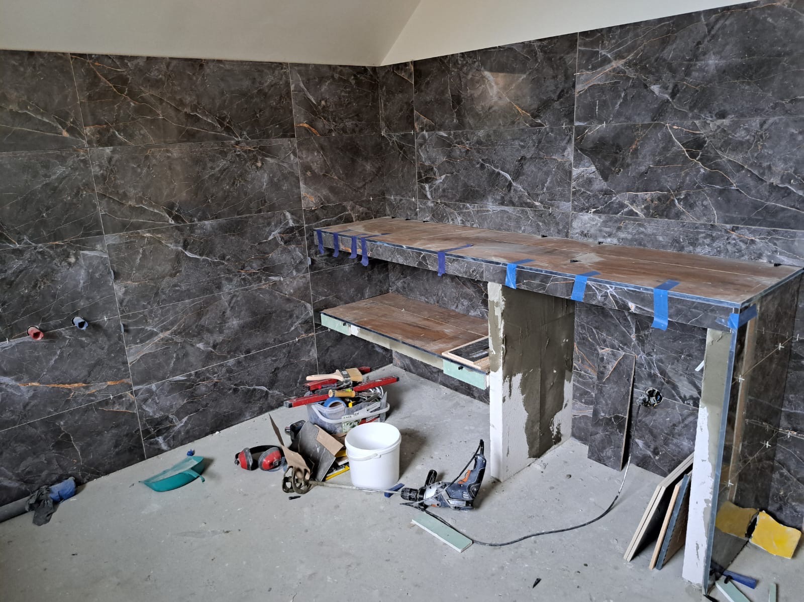 Montaż płytek imitujących marmur na ścianie i konstrukcji w łazience. Widoczne narzędzia glazurnicze i poziomica. Etap prac wykończeniowych przed fugowaniem.