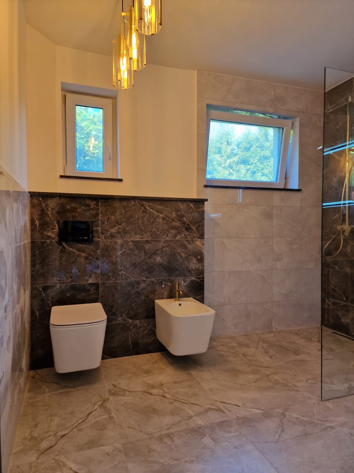 Elegancka łazienka z marmurowymi płytkami, wiszącą toaletą i bidetem. Dwa okna wpuszczają naturalne światło. Złota armatura dodaje luksusowego akcentu.
